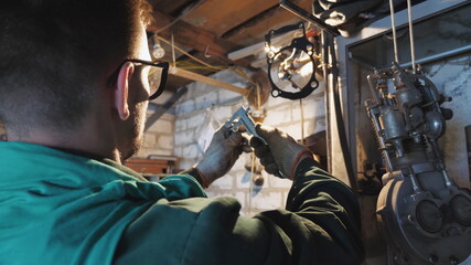 Male mechanic in protective glasses examines vehicle detail in garage. Male worker using tool for his work at workplace. Young repairman in uniform working in workshop. Close up Slow motion