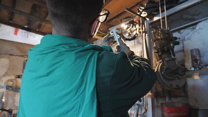 Male mechanic in protective glasses examines vehicle detail in garage. Male worker using tool for his work at workplace. Young repairman in uniform working in workshop. Close up Slow motion