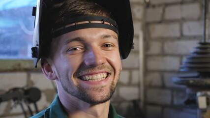 Close up of happy young welder opens protective mask after finishing his job. Portrait of mechanic in workwear smiling and looking into camera. Repairman with beard working in workshop. Dolly shot
