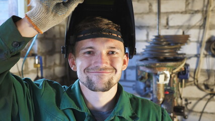 Portrait of mechanic in uniform making funny face and fooling around into camera. Happy young repairman showing grimaces and lowering protective mask in garage. Welder with beard working in workshop