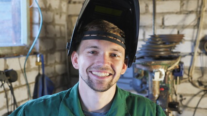Close up of happy young repairman opens protective mask after finishing his job. Portrait of mechanic in workwear smiling and looking into camera. Welder with beard working at garage. Slow motion