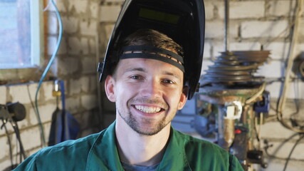 Close up of happy young repairman opens protective mask after finishing his job. Portrait of mechanic in workwear smiling and looking into camera. Welder with beard working at garage. Slow motion - Powered by Adobe