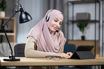 Remotely work from home concept. Close up of attractive smiling confident young arabic woman in headscarf, wearing headset for working from home on the tablet pc