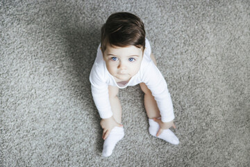 Infant child baby toddler kid in white bodysuit sitting on carpet and looking at the camera