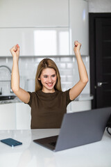 Happy smiling woman with win gesture sitting at table in kitchen drinking morning coffee reading message looking at device screen using computer.