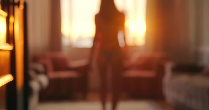 Young woman in lingerie walks towards the window in the apartment at sunset