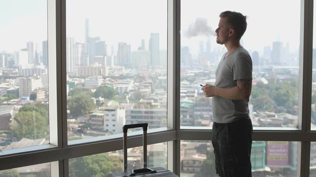 A man is enjoying smoking with wape in front of panoramic window with a luggage