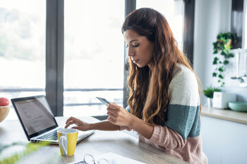 Cute young woman holding white credit card for shopping online with computer while sitting in the kitchen at home.