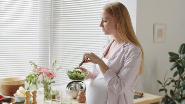 Tilt down shot of young Caucasian woman with big pregnant belly standing in kitchen and eating vegetable salad