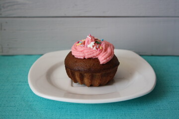 A small shaped cupcake with pink cream and sprinkles on a white plate.