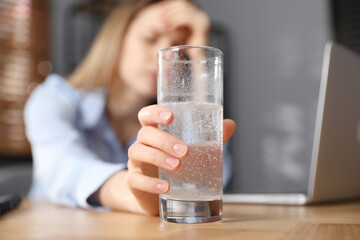 Woman taking medicine for hangover in office, focus on hand with glass