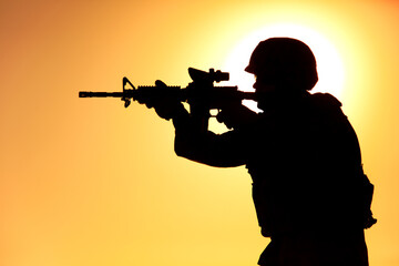 Silhouette of modern army infantry soldier standing on background of setting sun and aiming assault...