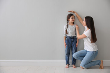Mother measuring little girl's height near light grey wall indoors. Space for text