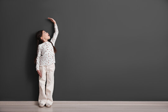 Emotional little child measuring her height near black wall indoors, space for text