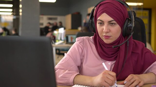 Happy smiling muslim student girl communicates online. Smiling islamic woman with headphones and a microphone having video call on laptop, talking and makes notes in a notebook. Multi-ethnic team