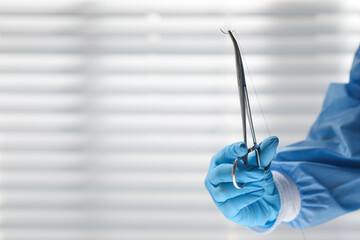 Doctor holding needle with suture thread indoors, closeup and space for text. Medical equipment