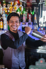 Smiling asian bartender shaking cocktail at the bar using metal mixer over colorful background with alcoholic beverages