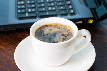 Coffee in a white cup and a laptop on the background on a brown wooden table. Concept of remote work or training, the start of the working day in the office.