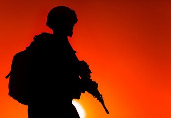 Silhouette of army rifleman in helmet and ammunition, carrying tactical backpack, walking with service rifle on background of sunset sky. Counter terrorist forces fighter marching at night mission