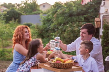 Happy family sits at a wooden table, drinks lemonade together by a trailer truck. Picnic, weekend camping with children. Travel in a motor home. Parents kiss, kids watch, laugh. Rest outside the city