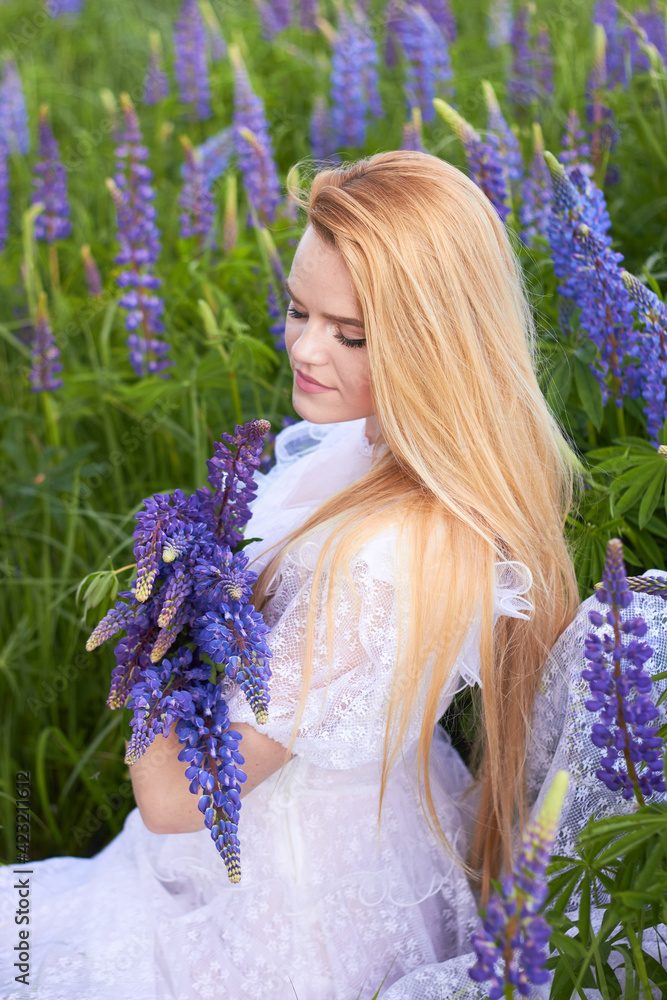 Wall mural young attractive long-haired blonde in a white vintage dress with a bouquet of lupins among a purple