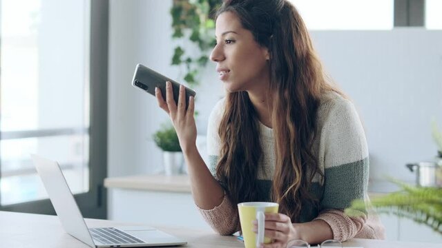 Video of concentrated young business woman using her mobile phone while working with laptop in the kitchen at home.