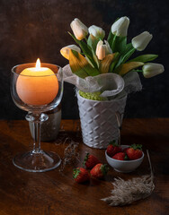 A round candle is lit in a glass candle holder near a vase of white flowers on a wooden table with some strawberries and dry plants