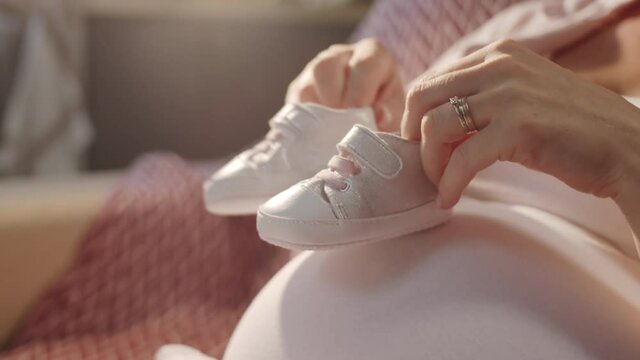 Close up shot of smiling expectant mother holding cute newborn shoes and making them walk on pregnant belly while waiting for baby