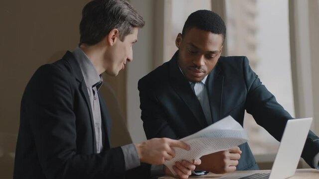 Two multiracial businessmen working on financial statistics analyzing project report doing paperwork sitting at office. Afro american executive discussing business diagrams wirh caucasian employee