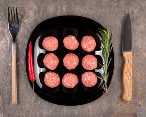 Raw meat balls on a black plate. Rosemary, chili pepper, knife, fork. Gray background.