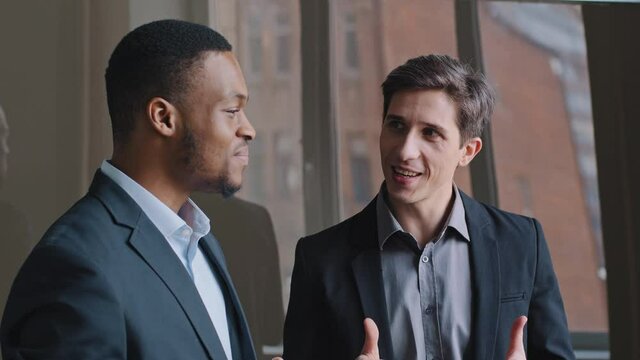 Two multiethnic multiracial men in formal suits couple businessmen african american man and caucasian employee stand near office window talking discussing argue planning laughing actively gesticulate