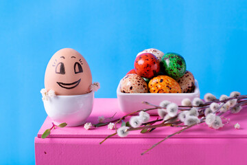 Funny Easter egg with cute face, colored candy eggs and willow twigs on wooden pink table against the blue background. Festive composition