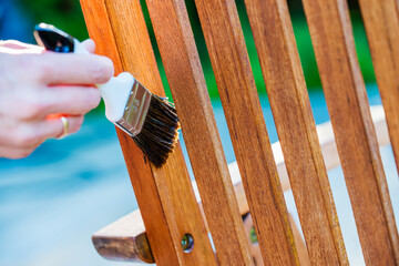 female hand holding a brush applying varnish paint on a wooden garden chair- painting and caring...
