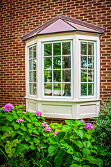 Vintage bay window with copper top reflecting leafy neighborhood -  pink hydrangea flower bed in foreground - red brick house