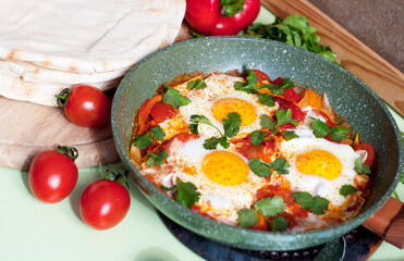 Traditional dish of israeli cuisine Shakshuka. Fried eggs with tomatoes, peppers, onions, cilantro and pita in a pan on light green background