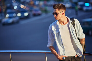 young man in sunglasses with a black backpack stands on the stairs leaning on the railing.