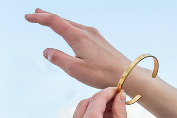 A girl measures a metal shiny bracelet as a piece of jewelry, putting it on her wrist against a blue sky background
