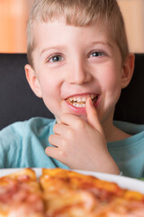 Beautiful happy young boy smiling and eating fresh made pizza,He sit at black chair, He has blonde hair.