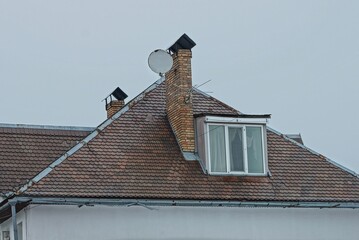 brown tiled roof of a private house with a large window, brick chimney and satellite dish against a...