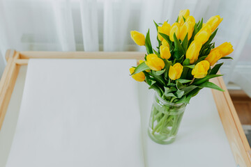 Fototapeta premium Bright fresh yellow tulips on white background with space for text. Bunch of yellow tulips in big glass jar in white interior. Spring flowers in glass vase on white table at home.