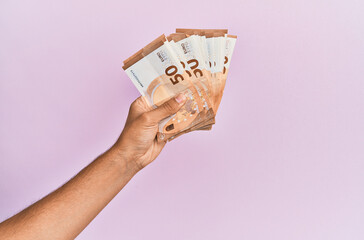 Hispanic hand holding 50 euro banknotes over isolated pink background.