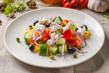 Greek salad on a large white plate on the table covered with a light linen cloth on a beautiful background of vegetables and herbs. Beautiful bokeh.