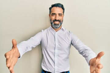 Middle age man with beard and grey hair wearing casual white shirt looking at the camera smiling with open arms for hug. cheerful expression embracing happiness.