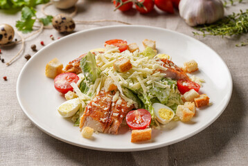 Caesar salad with chicken on a large white plate on the table covered with a light linen cloth on a beautiful background of vegetables and herbs. Beautiful bokeh.