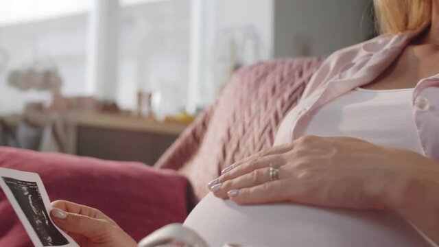 Close up shot of happy expectant mother sitting at home with bootees on lap, looking at fetal ultrasound image, stroking pregnant belly and smiling
