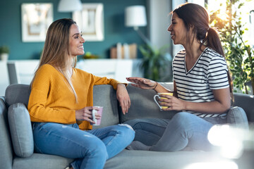 Two smiling young women talking while watching smartphone sitting on couch in the living room at home.
