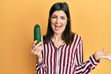 Young hispanic woman holding cucumber celebrating achievement with happy smile and winner expression with raised hand