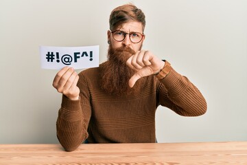Young irish redhead man holding paper with insult message sititng on the table with angry face,...
