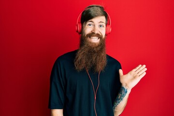 Redhead man with long beard listening to music using headphones smiling cheerful presenting and pointing with palm of hand looking at the camera.