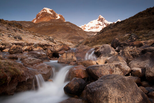 A long exposure of a creek rushing through boulders during sunrise over Mt Trapecio to the left and Jurau to the right located in the Cordillera Huayhuash of the Andes Mountains in Peru.     